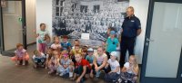 Zdjęcie grupowe policjanta i dzieci na tle plakatu przedstawiającego początki służb mundurowych w Dąbrowie Górniczej