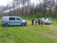 2 umundurowanych policjantów oraz 2 umundurowanych strażników leśnych, radiowóz oznakowany Poicji, pojazd oznakowany Straży Leśnej, na tle lasu.