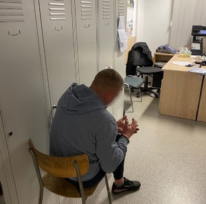 Zdjęcie przedstawia zatrzymanego siedzącego w pokoju przesłuchań. mężczyzna ma założone kajdanki na ręce trzymane z przodu.