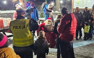 Umundurowani Policjanci i Sznupek wraz ze Świętym Mikołajem wręczają prezenty dzieciom.
