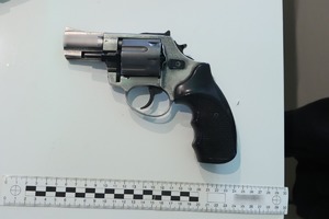 Pistolet koloru srebrno-czarnego, leżący na białym podłożu pod pistoletem linijka do stwierdzenia wielkości broni.