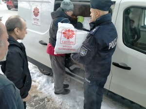 Umundurowani policjant i pracownik Caritas wręczają paczkę bezdomnym.